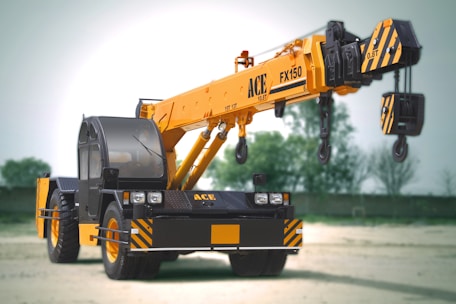 A large mobile crane with a yellow telescopic boom and black cab is positioned in an open area. It features the brand and model number ACE FX150 on its arm. The crane has black and yellow hazard striping on various parts, including weights and wheels, and stands on a dirt or sand surface with soft-focus green trees in the background.