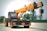 A large mobile crane with a yellow telescopic boom and black cab is positioned in an open area. It features the brand and model number ACE FX150 on its arm. The crane has black and yellow hazard striping on various parts, including weights and wheels, and stands on a dirt or sand surface with soft-focus green trees in the background.