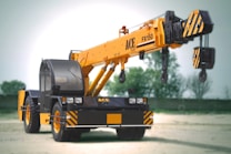 A large mobile crane with a yellow telescopic boom and black cab is positioned in an open area. It features the brand and model number ACE FX150 on its arm. The crane has black and yellow hazard striping on various parts, including weights and wheels, and stands on a dirt or sand surface with soft-focus green trees in the background.