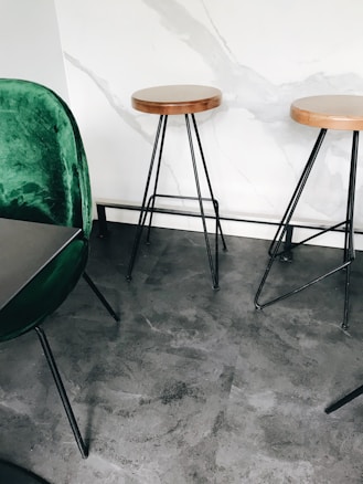 A modern interior space features two round wooden stools with black metal legs, positioned against a white marbled wall. A green velvet chair with black metal legs is partially visible on the left foreground. The floor appears to be made of polished concrete, contributing to a minimalist aesthetic.