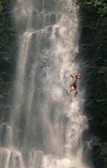 A person is mid-air in front of a large, cascading waterfall. The individual appears to be wearing red shorts and is joyfully posing with arms flexed. The scene conveys a sense of adventure and freedom, surrounded by greenery and mist from the waterfall.