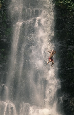 A person is mid-air in front of a large, cascading waterfall. The individual appears to be wearing red shorts and is joyfully posing with arms flexed. The scene conveys a sense of adventure and freedom, surrounded by greenery and mist from the waterfall.