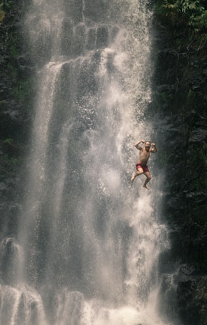 A person is mid-air in front of a large, cascading waterfall. The individual appears to be wearing red shorts and is joyfully posing with arms flexed. The scene conveys a sense of adventure and freedom, surrounded by greenery and mist from the waterfall.