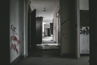 A dimly lit hallway in an abandoned building, with doors slightly ajar and graffiti on the walls. The floor is covered with debris, and the atmosphere is eerie and desolate.