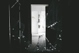 A dark interior space with a shattered glass window in the foreground. Light from an open door in the background illuminates part of the room. The open door reveals a bright outdoor area and displays some graffiti on the wall beside it.