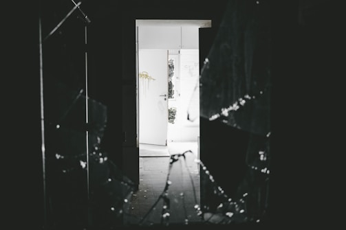 A dark interior space with a shattered glass window in the foreground. Light from an open door in the background illuminates part of the room. The open door reveals a bright outdoor area and displays some graffiti on the wall beside it.