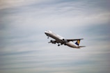 A commercial airplane is in flight, ascending into the sky. The aircraft is painted white with some visible branding on the side and tail. The background consists of a vast expanse of cloudy sky, indicating an overcast day.
