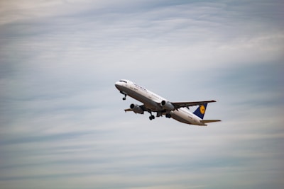 A commercial airplane is in flight, ascending into the sky. The aircraft is painted white with some visible branding on the side and tail. The background consists of a vast expanse of cloudy sky, indicating an overcast day.