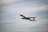 A commercial airplane is in flight, ascending into the sky. The aircraft is painted white with some visible branding on the side and tail. The background consists of a vast expanse of cloudy sky, indicating an overcast day.