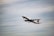 A commercial airplane is in flight, ascending into the sky. The aircraft is painted white with some visible branding on the side and tail. The background consists of a vast expanse of cloudy sky, indicating an overcast day.