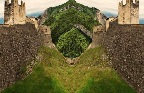A stone fortress with tall towers ascends from a lush green hill surrounded by steep mountains. The structure's architecture appears ancient, with weathered stone walls and crenelated battlements.