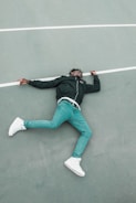 A person lies on their back on a sports court, with one arm slightly bent and the other extended. They are wearing a black jacket, blue jeans, and white sneakers. The ground is a muted grayish-green with a white line running horizontally.