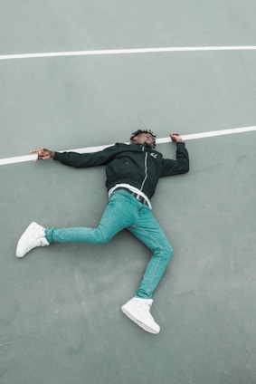 A person lies on their back on a sports court, with one arm slightly bent and the other extended. They are wearing a black jacket, blue jeans, and white sneakers. The ground is a muted grayish-green with a white line running horizontally.