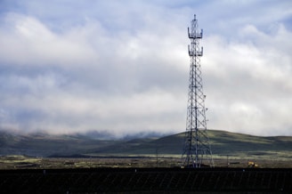 A tall communication tower stands prominently against a backdrop of rolling green hills and a cloudy sky. The landscape is expansive, with patches of sunlight breaking through the clouds, casting soft shadows on the ground.