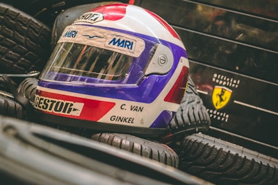 A colorful racing helmet with red, white, and blue colors is placed on a stack of tires. The name C. Van Ginkel is visible on the helmet. In the background, there is a logo resembling a Ferrari symbol on a black surface.