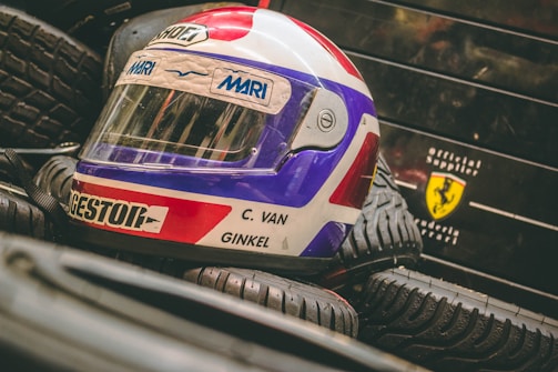 A colorful racing helmet with red, white, and blue colors is placed on a stack of tires. The name C. Van Ginkel is visible on the helmet. In the background, there is a logo resembling a Ferrari symbol on a black surface.