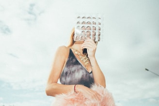 A person is standing outdoors, holding a sheet of bubble wrap in front of their face, obscuring their features. They are wearing a sleeveless outfit with a gold statement necklace. The background is a cloudy sky, and the person is holding a fuzzy pink object in their other hand.