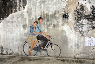 A mural on a weathered wall depicts a man and a woman riding a bicycle. The couple is artfully painted to appear as if they are sitting on a real bicycle, creating a playful and three-dimensional effect. The wall's texture adds character and depth to the artwork, with areas of peeling paint and discoloration surrounding the painting. A piece of paper with text is pasted to the right side of the image.
