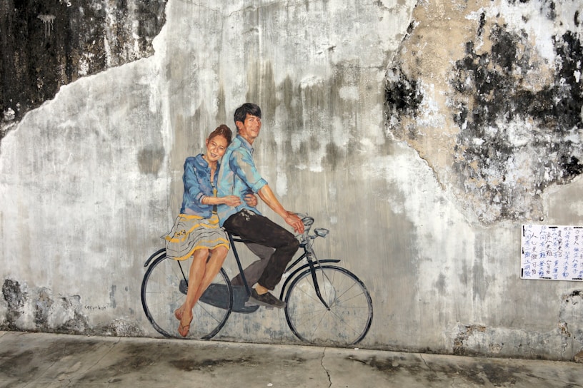 A mural on a weathered wall depicts a man and a woman riding a bicycle. The couple is artfully painted to appear as if they are sitting on a real bicycle, creating a playful and three-dimensional effect. The wall's texture adds character and depth to the artwork, with areas of peeling paint and discoloration surrounding the painting. A piece of paper with text is pasted to the right side of the image.