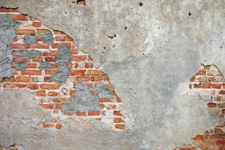 A weathered brick wall partially covered in deteriorating plaster. The red bricks are exposed in several areas, while gray plaster is peeling and rough in texture.