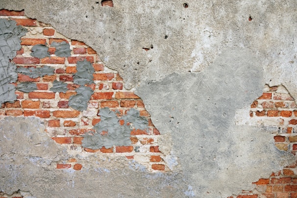 A weathered brick wall partially covered in deteriorating plaster. The red bricks are exposed in several areas, while gray plaster is peeling and rough in texture.