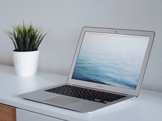 A sleek silver laptop sits on a clean, white desk. The screen displays a calming image of water with gentle waves. To the left of the laptop, a small green potted plant adds a touch of nature to the minimalist setting. The background is a plain, light-colored wall.