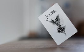 A playing card featuring a joker design is in mid-air against a blurred background. The card displays an illustration of a winged figure standing over another figure. The card's text reads 'JOKER' in bold letters at the top.