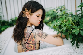 A woman with long, dark wavy hair is seen with an intricate tattoo on her upper back, sitting amidst green plants. Her hair is adorned with a delicate, leaf-shaped hair accessory. The setting is serene with white stones and lush foliage surrounding her.