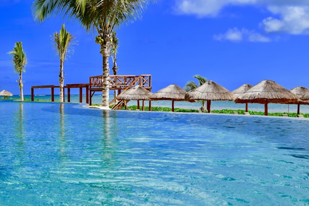 A tranquil scene of a tropical resort features an infinity pool stretching towards the horizon. Palm trees are scattered around the area, beside wooden structures and thatched umbrellas lining the edge of the pool. The sky is vividly blue, complementing the turquoise water, creating a serene and idyllic atmosphere.