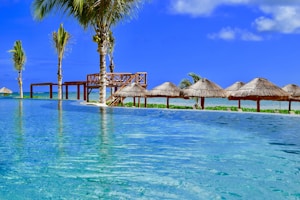 A tranquil scene of a tropical resort features an infinity pool stretching towards the horizon. Palm trees are scattered around the area, beside wooden structures and thatched umbrellas lining the edge of the pool. The sky is vividly blue, complementing the turquoise water, creating a serene and idyllic atmosphere.