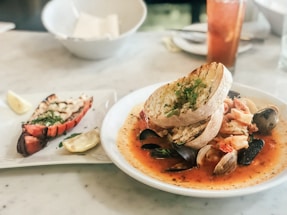 A seafood dish is presented on a plate, featuring clams, mussels, and pieces of shrimp in a tomato-based broth, garnished with herbs. Toasted slices of bread are placed on top of the dish. Next to it, there is a plate with a grilled lobster tail garnished with herbs and a wedge of lemon. A glass of iced tea is visible in the background.