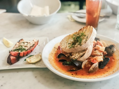 A seafood dish is presented on a plate, featuring clams, mussels, and pieces of shrimp in a tomato-based broth, garnished with herbs. Toasted slices of bread are placed on top of the dish. Next to it, there is a plate with a grilled lobster tail garnished with herbs and a wedge of lemon. A glass of iced tea is visible in the background.