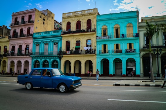 A vibrant street scene features a row of colorful colonial-style buildings in pastel pink, yellow, and turquoise. A vintage blue car drives along an empty road, with a few pedestrians walking on the sidewalk. The sky is mostly clear with some white clouds, and there is a palm tree on the right.