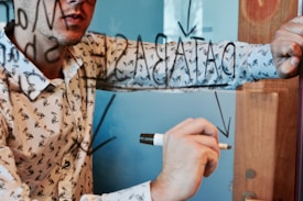 A person wearing a patterned shirt writes on a glass surface with a marker. Reflections are visible on the transparent surface, and some words are partly obscured. The background has a soft blue tone.