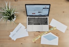 A wooden desk features a laptop displaying an email marketing webpage. On the desk are envelopes, one open with a blue inner lining, and two visible notes with the phrase 'Email Marketing.' There is also a small potted plant with green leaves and a yellow tape labeled 'Saturday Delivery.'