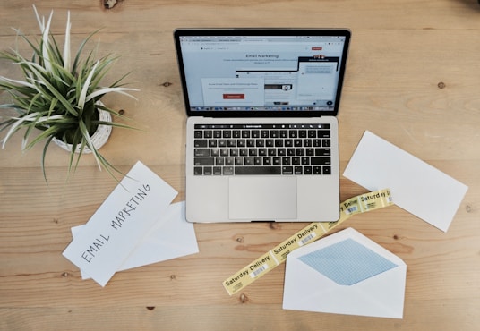 A wooden desk features a laptop displaying an email marketing webpage. On the desk are envelopes, one open with a blue inner lining, and two visible notes with the phrase 'Email Marketing.' There is also a small potted plant with green leaves and a yellow tape labeled 'Saturday Delivery.'