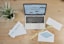 A wooden desk features a laptop displaying an email marketing webpage. On the desk are envelopes, one open with a blue inner lining, and two visible notes with the phrase 'Email Marketing.' There is also a small potted plant with green leaves and a yellow tape labeled 'Saturday Delivery.'