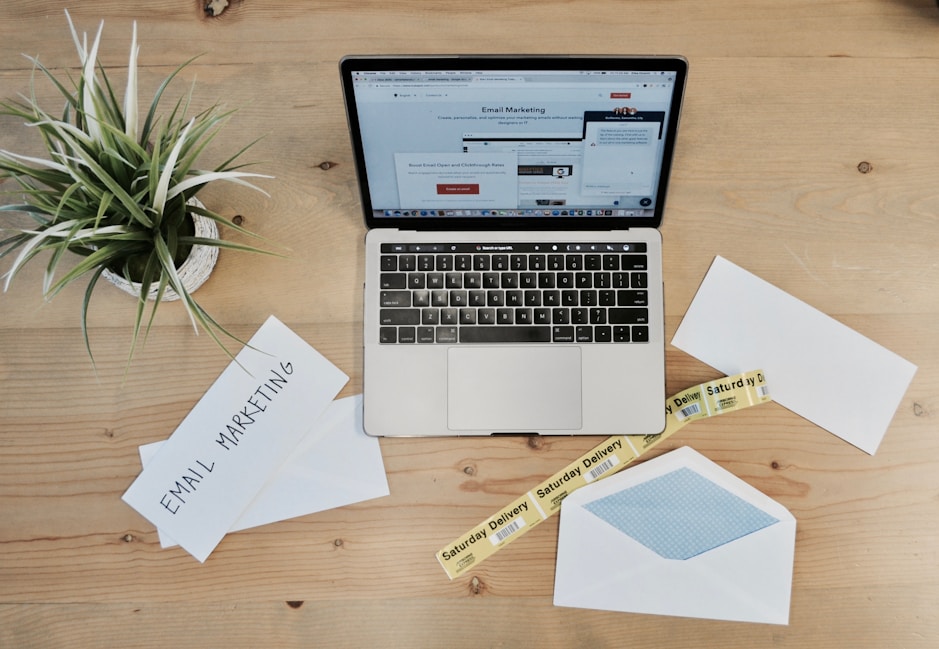 A wooden desk features a laptop displaying an email marketing webpage. On the desk are envelopes, one open with a blue inner lining, and two visible notes with the phrase 'Email Marketing.' There is also a small potted plant with green leaves and a yellow tape labeled 'Saturday Delivery.'