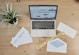 A wooden desk features a laptop displaying an email marketing webpage. On the desk are envelopes, one open with a blue inner lining, and two visible notes with the phrase 'Email Marketing.' There is also a small potted plant with green leaves and a yellow tape labeled 'Saturday Delivery.'