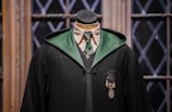 A mannequin dressed in a black academic-style robe with a green lining is positioned against a backdrop of wooden window frames with diamond-patterned glass. The attire includes a white shirt, a green striped tie, and a gray sweater vest beneath the robe. A decorative emblem featuring a shield-like design is pinned on the chest.