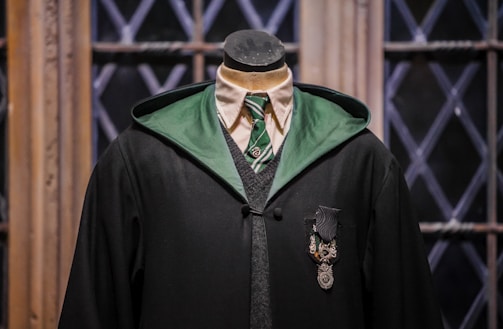 A mannequin dressed in a black academic-style robe with a green lining is positioned against a backdrop of wooden window frames with diamond-patterned glass. The attire includes a white shirt, a green striped tie, and a gray sweater vest beneath the robe. A decorative emblem featuring a shield-like design is pinned on the chest.