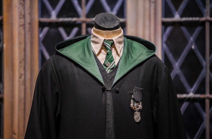 A mannequin dressed in a black academic-style robe with a green lining is positioned against a backdrop of wooden window frames with diamond-patterned glass. The attire includes a white shirt, a green striped tie, and a gray sweater vest beneath the robe. A decorative emblem featuring a shield-like design is pinned on the chest.
