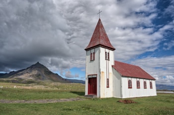 A small, rustic church with a white facade and red roof sits in an expansive green field. The church features a tall steeple topped with a cross. In the background, a rugged mountain rises under a partly cloudy sky.