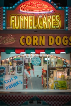 A brightly lit food stand at a fair or carnival features neon signs advertising funnel cakes and corn dogs. The booth is adorned with colorful lights and signs for cold drinks, lemonade shake-ups, and bottled water. Inside, a person is preparing or serving food surrounded by various supplies and equipment.