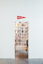 A well-organized bookshelf filled with books is visible through an open doorway in a room with a shiny, polished floor. Above the doorway, a red triangular pennant with the word 'READ' is displayed. Various books, both vertically and horizontally stacked, line the shelves, and a small part of a wheeled cart is visible.