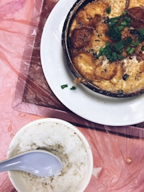 A cooked dish in a black skillet garnished with chopped green onions sitting on a white plate. Next to it is a bowl of plain white rice with a gray spoon. The table is covered with a pink plastic tablecloth.