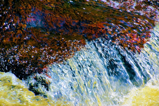 Water flows over a rocky surface, creating a small waterfall effect. The water appears clear and dynamic, cascading over textured rocks with hues of brown, orange, and reddish tones.