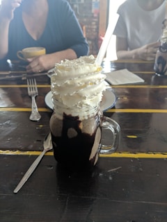 A creamy beverage topped with generous swirls of whipped cream, served in a glass jar with chocolate drizzles. The jar has a handle and a straw inserted into it. In the background, people are partially visible, sitting at a rustic wooden table with other items like a cup of coffee, a fork, and a napkin.
