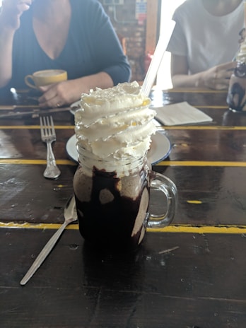 A creamy beverage topped with generous swirls of whipped cream, served in a glass jar with chocolate drizzles. The jar has a handle and a straw inserted into it. In the background, people are partially visible, sitting at a rustic wooden table with other items like a cup of coffee, a fork, and a napkin.