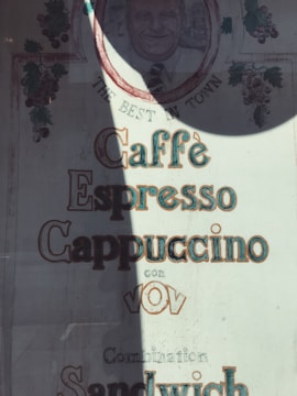 A vintage-style sign features a smiling man's illustration and text advertising Caffè Espresso Cappuccino. Vines and grapes decorate the borders. The words 'The Best in Town' are above the illustration.